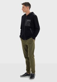 Felpa nera con zip e tasca frontale testurizzata, abbinata a pantaloni verde oliva e sneakers grigie, con un design semplice.