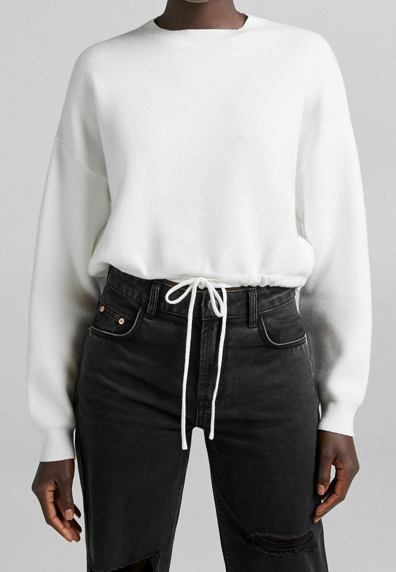 Personne portant un pull en tricot blanc à manches longues avec une taille à cordon et un jean noir déchiré taille haute.