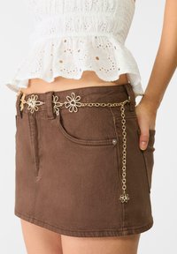 Mini-jupe en denim marron avec poches avant, ornée d'une ceinture en chaîne dorée embellie de motifs floraux et d'une fermeture à bouton à la taille.