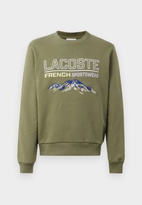 Felpa color verde oliva con maniche lunghe, collo a giro, con la scritta "LACOSTE FRENCH SPORTSWEAR" e un design di montagna ricamato in blu.