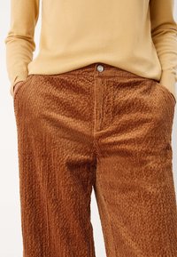 Pantalon en velours côtelé marron avec des côtes verticales, doté d'une fermeture à bouton à l'avant et de poches latérales, associé à un pull ajusté jaune clair.