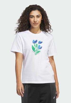 Femme aux cheveux bouclés portant un t-shirt blanc avec un motif floral bleu et vert et un pantalon Adidas noir, debout devant un fond uni.
