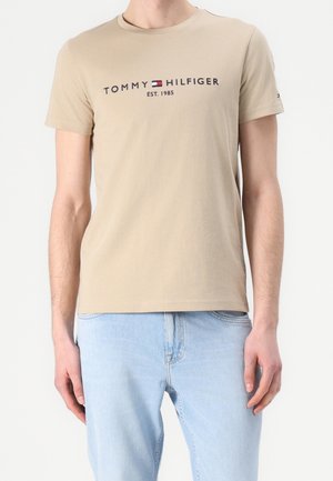 Beige Tommy Hilfiger T-Shirt mit Logo und dem Schriftzug „EST. 1985“, getragen von einer Person mit hellblauen Jeans, die vor einem einfarbigen Hintergrund steht.