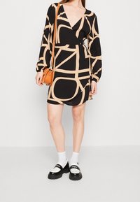 Schwarzes Kleid mit beigem abstraktem Muster, V-Ausschnitt und langen Puffärmeln. Das Model trägt schwarze Loafers mit weißen Akzenten und hält eine kleine orangefarbene Tasche.