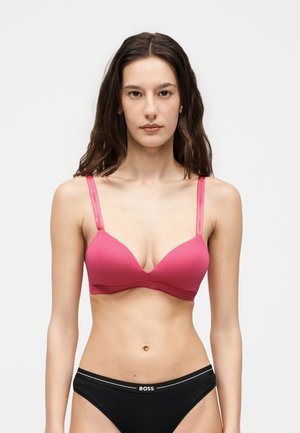 Kvinde med langt brunt hår iført en pink bh og sort BOSS undertøj, stående mod en ensfarvet lys baggrund, vendt fremad.