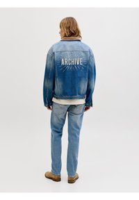 Άτομο με την πλάτη γυρισμένη φοράει μια ξεθωριασμένη μπλε τζιν ζακέτα με το κείμενο «ARCHIVE» και ακτίνες, σε συνδυασμό με ανοιχτό μπλε τζιν παντελόνι και καφέ μπότες.