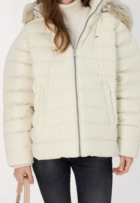 Dynejakke i creme med lynlås foran, vandret syning og aftagelig hætte foret med pels. Har sidelommer og ribstrikkede manchetter.