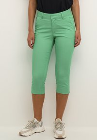 Green cropped pants in a stretchy fabric, featuring a smooth texture and a fitted design. Paired with light sneakers for a casual look.