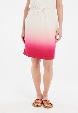 Vrouw draagt een knielange rok met roze naar witte ombré, een taille met trekkoord en bruine leren sandalen, staand op een witte achtergrond.