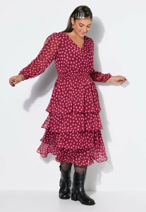 Vrouw die lacht en omlaag kijkt, draagt een lang mouwige gelaagde roze polkadotjurk met zwarte laarzen en haarclips tegen een witte achtergrond.