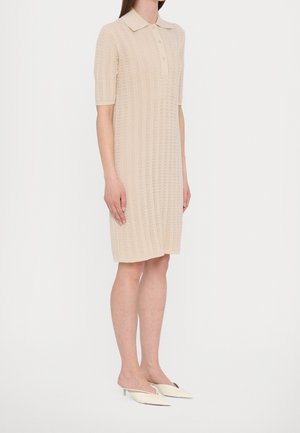 Femme portant une robe en maille beige mi-longue avec un col et des manches courtes, associée à des mules à talons crème à bouts pointus.