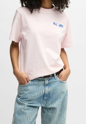 Person iført en lyserød t-shirt med et lille blåt og pink blomsterdesign, kombineret med lyseblå jeans med hvide skriftmønstre.