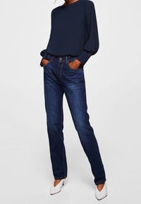 Blouse bleu marine à manches longues bouffantes, associée à un jean droit bleu foncé et des escarpins blancs pointus, posée sur un fond neutre.