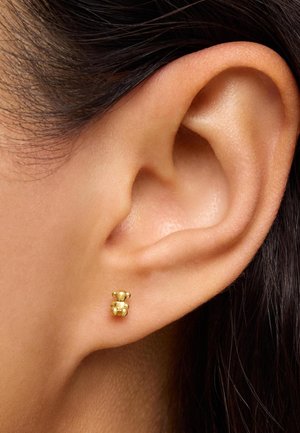Pendiente en forma de osito de oro con formas redondeadas y acabado pulido, mostrado en el lóbulo de una oreja con cabello oscuro cerca.