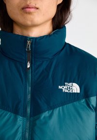 Turbia i niebieska pikowana kurtka z wysokim kołnierzem, z zamkiem błyskawicznym z przodu oraz haftowanym na białe logo "The North Face" na klatce piersiowej.