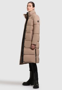 Beige puffer-coat, jossa on korkea kaulus, vetoketju ja nappikiinnitys, sivutaskut sekä pitkä, toppattu muotoilu. Yhdistetty mustiin saappaisiin.