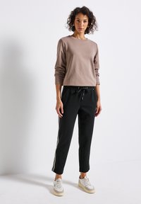 Kvinna som står klädd i en ljusbrun långärmad överdel, svarta byxor med dragsko och vita sidosträck, samt vita sneakers mot en enfärgad vit bakgrund.