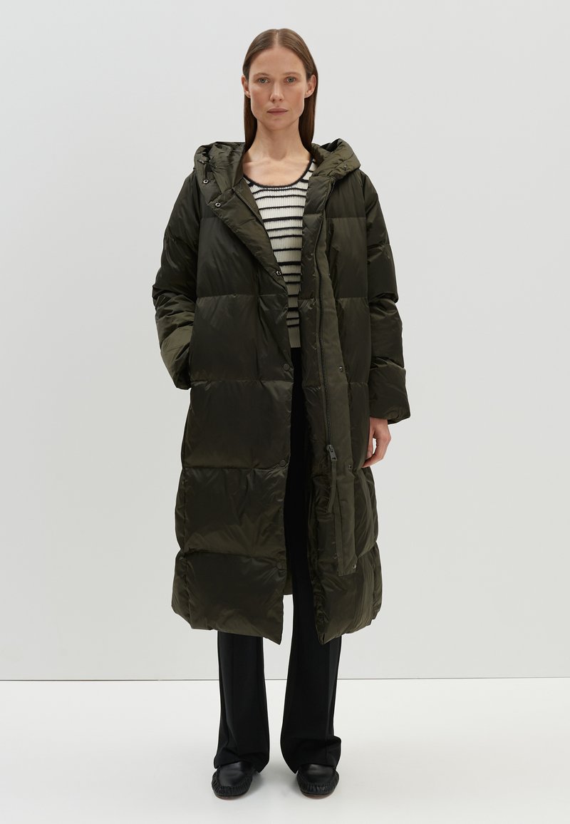 Lange olijfgroene pufferjas met capuchon, voorzien van een ritssluiting, drukknopen en een gestikt ontwerp. Gepaard met een zwart-wit gestreept shirt.