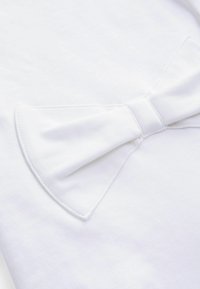 Indumento in cotone bianco con un evidente dettaglio a fiocco, tessuto strutturato e cuciture pulite, che enfatizzano una superficie liscia e uniforme.
