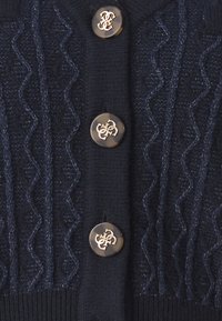 Tmavě modrý pletený cardigan s texturovaným vlnovitým vzorem a zlatými logovými knoflíky. Žebrovaný lem a manžety dodávají designu detail.