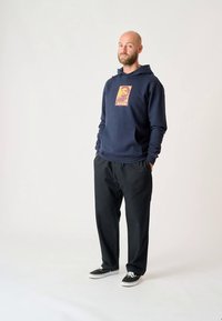 Námořnická mikina s kapucí s grafickým motivem "AHOI" na hrudi, doplněná černými kalhotami a černými botami. Jednoduchý, ležérní outfit.