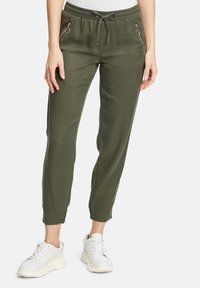Olive green jogger pants with an elastic waistband and drawstring, featuring zippered side pockets and tapered ankles. Paired with white sneakers.