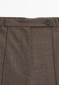 Gros plan de la ceinture d’un pantalon en laine marron avec dos élastique et un seul bouton noir sur le côté droit.