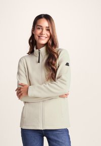 Light beige fleece jacket with a high collar, front zipper, and side pockets; features a black logo patch on the left sleeve.