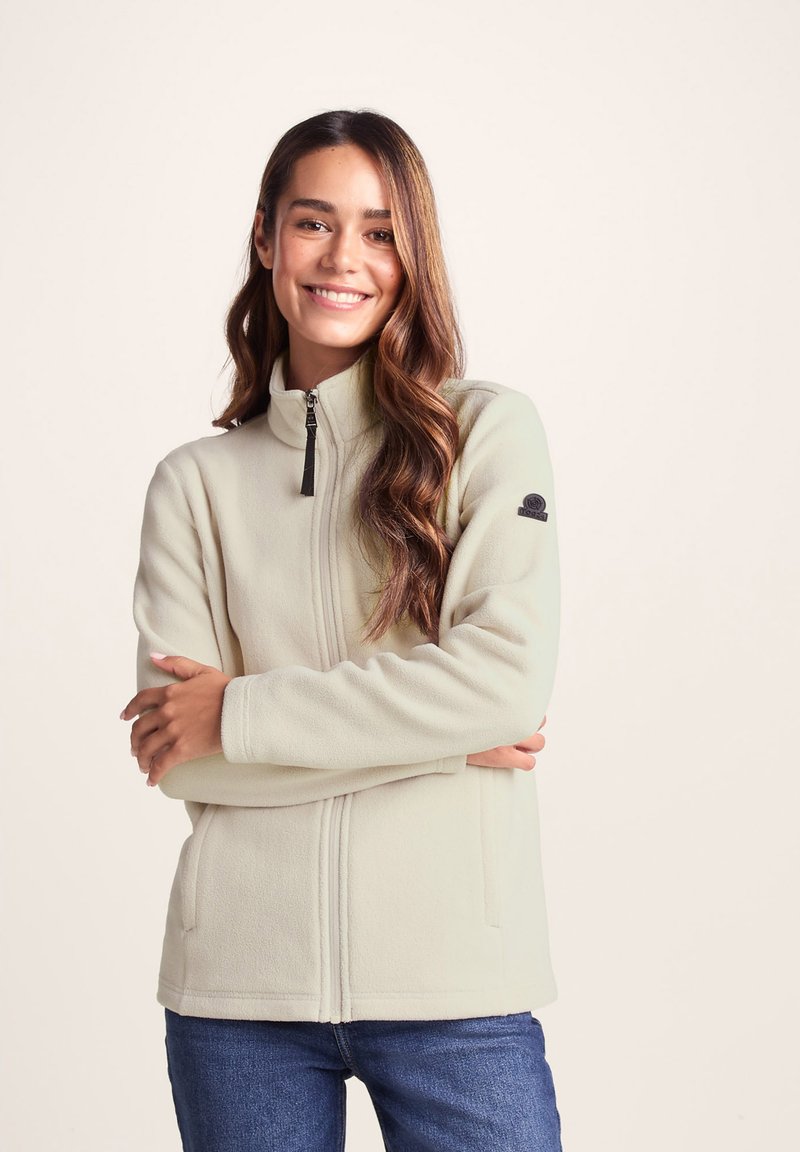 Light beige fleece jacket with a high collar, front zipper, and side pockets; features a black logo patch on the left sleeve.