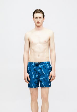 Νέος άνδρας με κοντά καστανά μαλλιά που φοράει μπλε μαγιό με σχέδιο Nike, στέκεται μπροστά σε απλό λευκό φόντο.