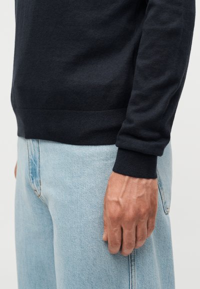 Αντρικός κορμός και χέρι με μαύρο πουλόβερ με μακριά μανίκια και ανοιχτό μπλε τζιν, σε απλό φόντο.