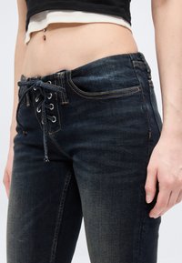 Μέση ενός ατόμου που φοράει σκούρα μπλε τζιν με κορδόνια και κοντό τοπ σε ασπρόμαυρο χρώμα, εμφανίζοντας μια τρύπα αφαλού με σκουλαρίκι.