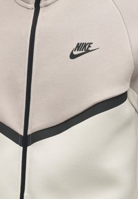 Μπεζ και ανοιχτό γκρι ζακέτα Nike με φερμουάρ σε μαύρο χρώμα και μαύρο λογότυπο Nike στο πάνω αριστερό μέρος του στήθους.