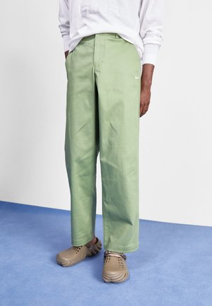 Person trägt hellgrüne, weit geschnittene Hosen und beige Slipper, steht auf blauem Boden vor einer weißen Wand.