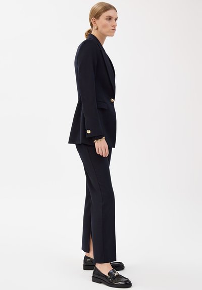 Femme portant un blazer noir cintré avec des boutons dorés et un pantalon noir assorti avec des fentes latérales, associé à des mocassins noirs et des bijoux dorés.