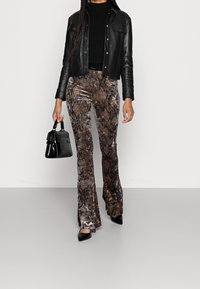 Nainen, jolla on yllään musta nahkatakki, musta toppi, ruskeat käärmeennahkakuosiset samettihousut, mustat kärkitossut ja joka pitää kädessään pientä mustaa käsilaukkua.