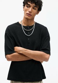 Chemise noire à manches courtes et à boutons, en tissu texturé et à coupe décontractée. Colliers superposés et bras croisés devant.
