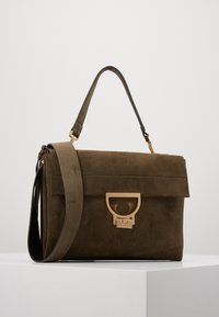 Brązowa zamszowa torba na ramię z przednim złotym zapięciem, odpinanym paskiem i górnym uchwytem, spoczywająca na białej powierzchni na jednolitym tle.