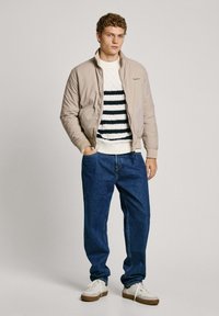 Beige bomberjacka över en vit tröja med svarta ränder, i kombination med blå jeans och vita sneakers. Enkelt design med en avslappnad passform.