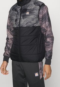 Vadderad väst med svart quiltad nederdel och grå marmorliknande mönster på övre delen, med hög krage och logotypmärke på bröstet.