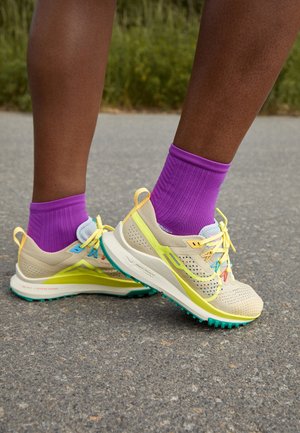 Jambes portant des chaussettes violettes et des baskets beiges avec des accents jaunes et verts, debout sur une surface pavée près d'un feuillage vert.