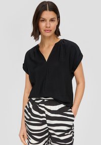Schwarze Bluse mit V-Ausschnitt, kurzen hochgeschlagenen Ärmeln und einer weichen, leichten Textur. Getragen mit schwarz-weiß gestreiften Hosen.