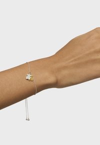 Pulsera de plata con un charm floral que presenta acentos en oro y plata, cadena delicada y longitud ajustable con cuentas al final.