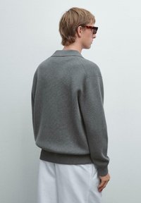 Grijze gebreide trui met een kraag, relaxte pasvorm, geribde manchetten en zoom, gedragen over witte broeken. De textuur lijkt zacht en knus.