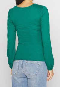 Donna che indossa un top verde attillato a maniche lunghe e jeans chiari a vita alta, vista da dietro contro uno sfondo semplice.