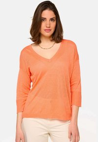 Femme aux cheveux bruns mi-longs portant un pull V en léger orange et un pantalon beige sur fond blanc.
