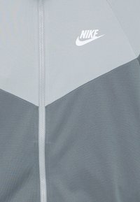 Γκρι Nike μπουφάν με άσπρο λογότυπο. Διαθέτει φερμουάρ και σχέδιο με χρώματα, που συνδυάζει ανοιχτές και σκούρες γκρι περιοχές.