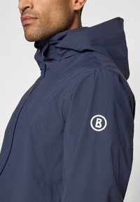 Navy waterproof jacket with a high collar and adjustable hood, featuring a circular logo on the left sleeve. Smooth texture, streamlined design.