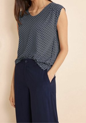 Frau trägt ärmelloses, marineblau gemustertes Oberteil, in hoch taillierte, weite marineblaue Hosen gesteckt, Hand in der Tasche vor einfarbigem Hintergrund.