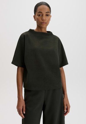 Donna con capelli scuri raccolti, indossa una maglia corta a coste scura e larga e pantaloni larghi scuri abbinati su sfondo uniforme.